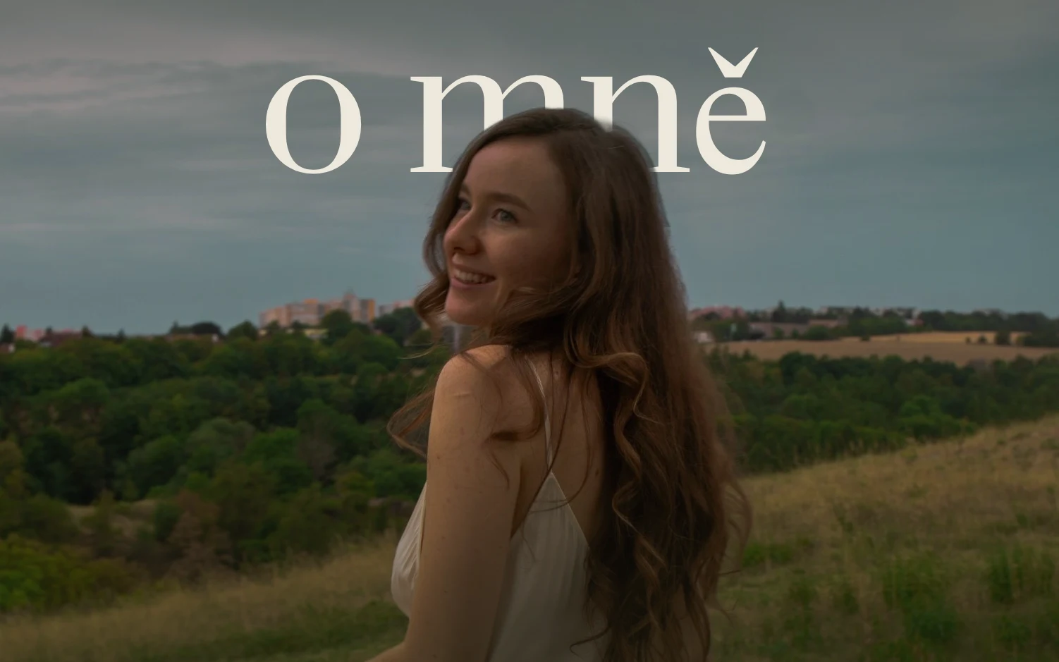 Aňa se ohlíží přes rameno a usmívá se, za ní pole a kopce