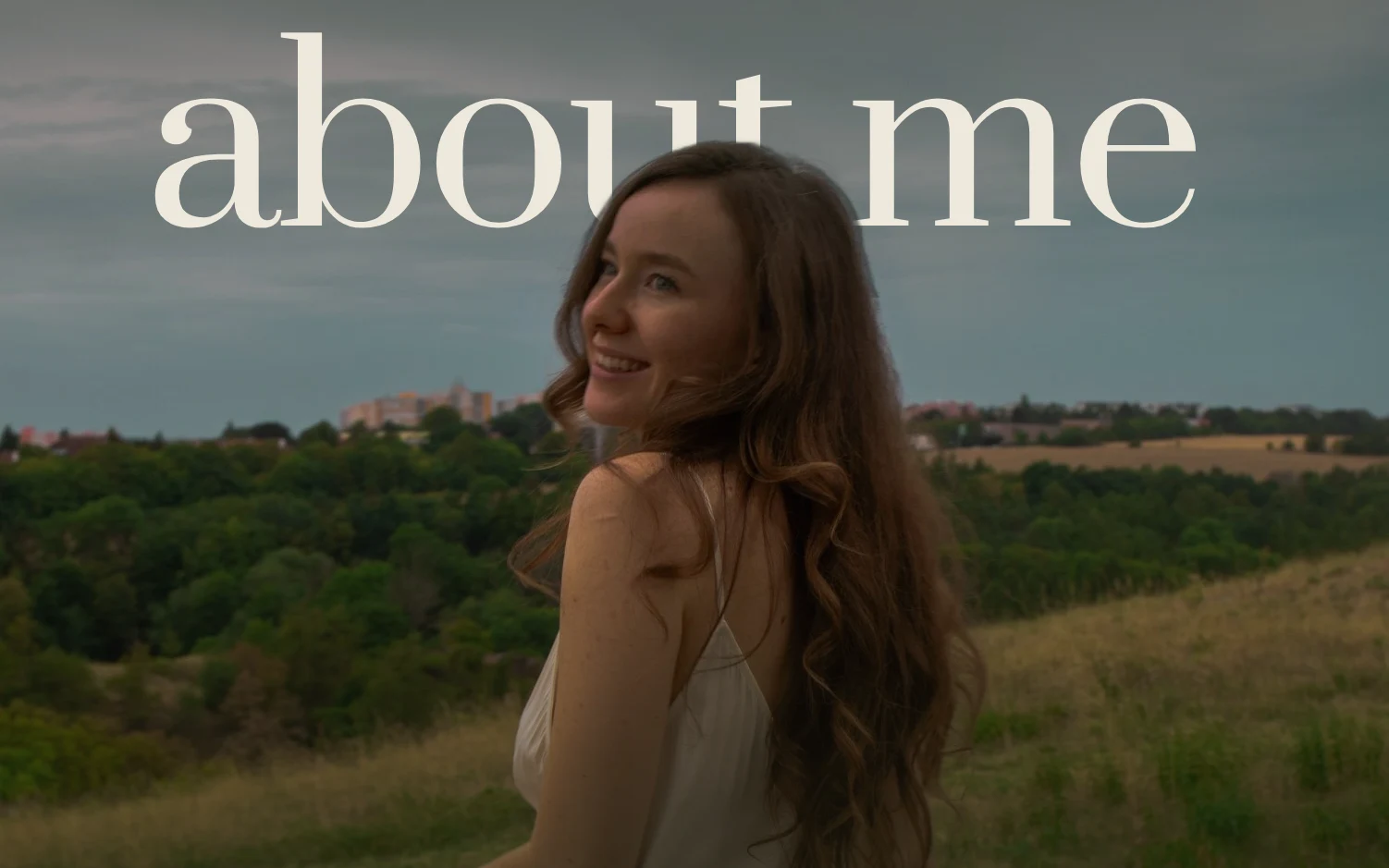 Anya looks back over her shoulder and smiles, a field and hills behind her