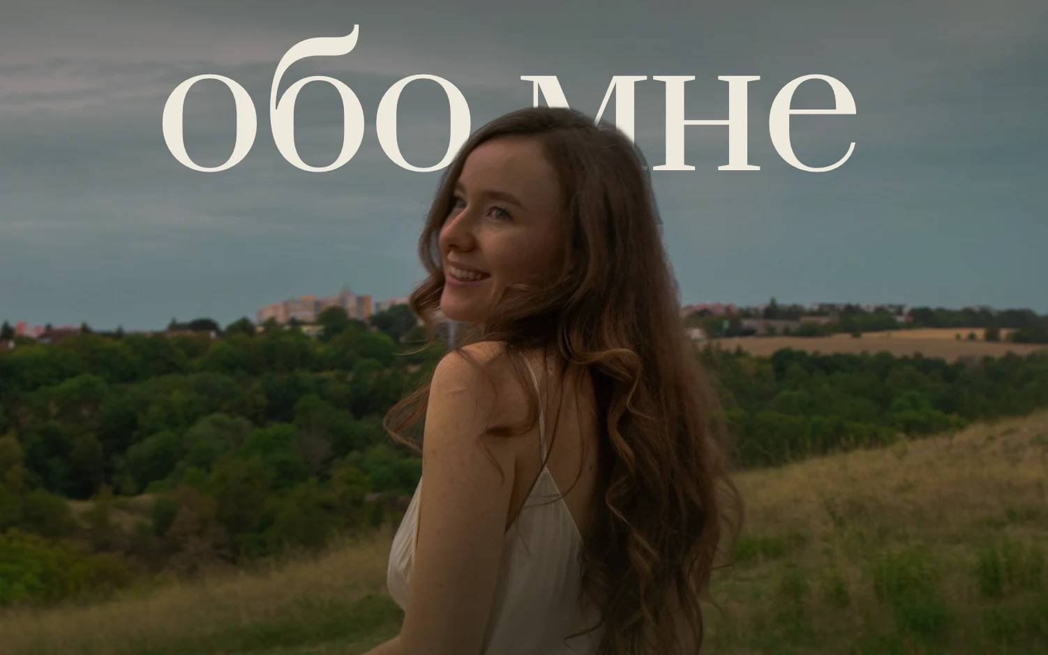 Аня оборачивается через плечо и улыбается, за ней поле и холмы